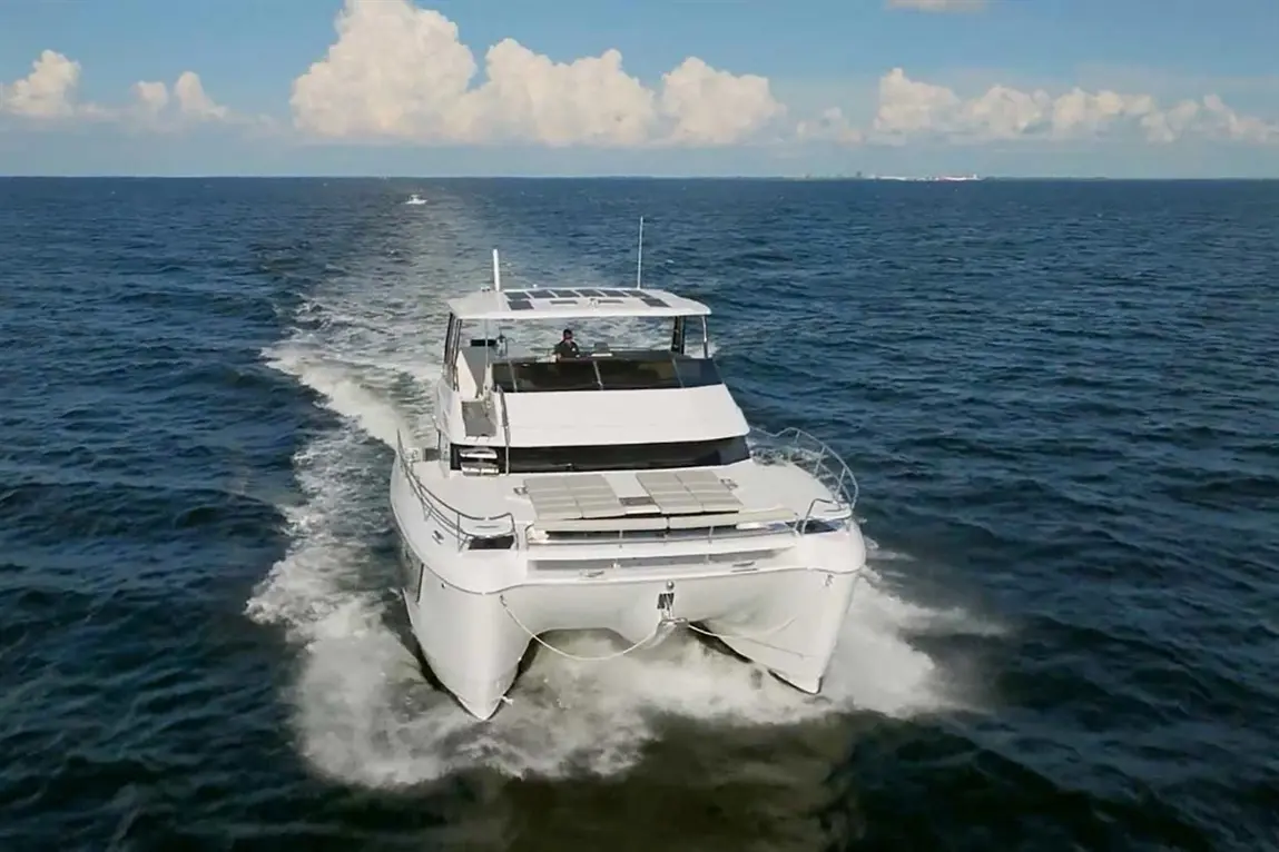 A white catamaran cruising on deep blue water, with fluffy clouds in the sky and a person steering at the helm.
